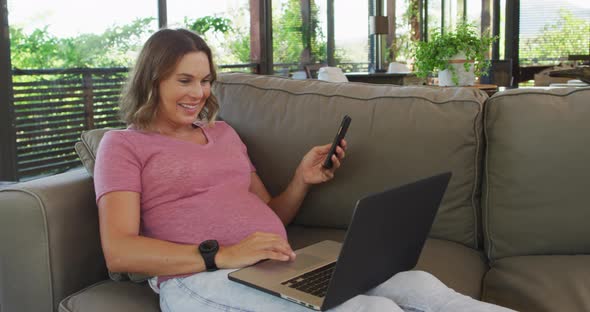 Happy caucasian pregnant woman sitting on sofa and using smartphone and laptop