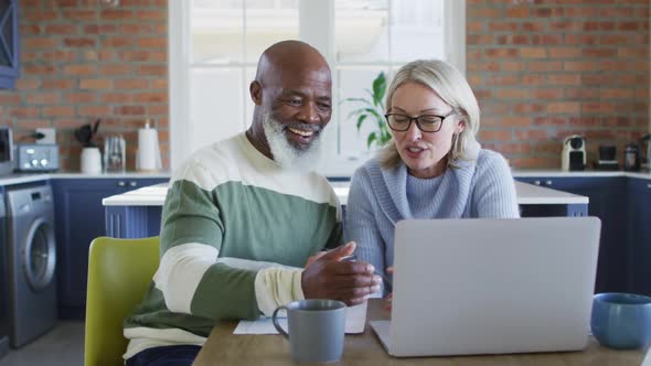 Happy senior diverse couple in kitchen sitting at table, using laptop