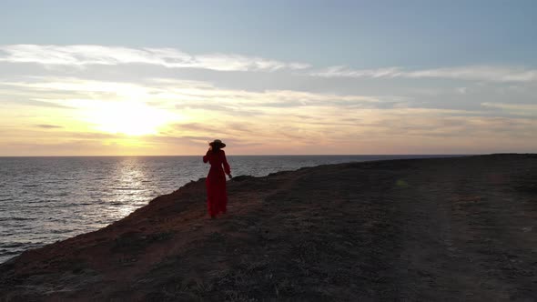 A Young Slender Woman in a Red Dress and Hat Walks Next to a Cliff of a Rocky Seashore at Sunset