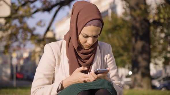 A Young Beautiful Muslim Woman Works on a Smartphone in a Park in an Urban Area  Closeup