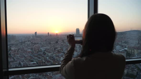 Woman Takes Photos of Panoramic City View at Sunset on Smartphone in Her Modern Flat in Skyscraper.