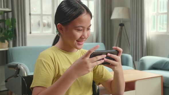 Excited Asian Kid Girl Sitting On Wheelchair Playing Video Game On Mobile Phone In House