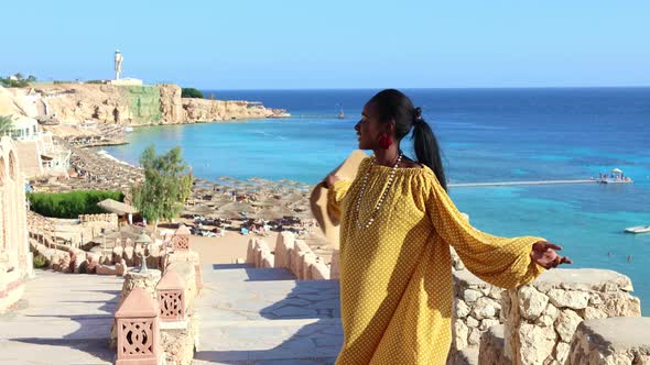 Happy African American Woman in Yellow Dress and Sun Hat Enjoys View of Coast of Red Sea on Natural