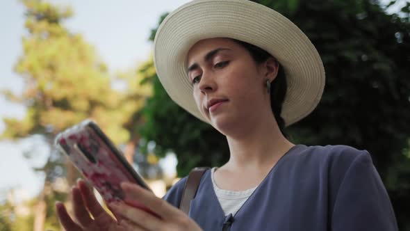 Portrait of a young woman in a straw hat standing on the street and typing on a smartphone.