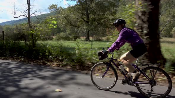 Side shot of a mature woman biking with her son along a shady rural road with a farm in the backgrou