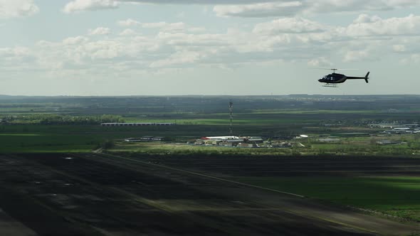 A Dark Helicopter Flies Against the Background of a Huge Blue Sky and Green Field on a Sunny Day