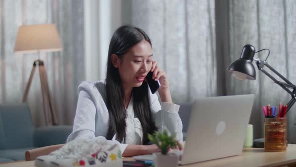 Asian Female Footwear Designer Talking On Smartphone While Working On A Laptop At Home