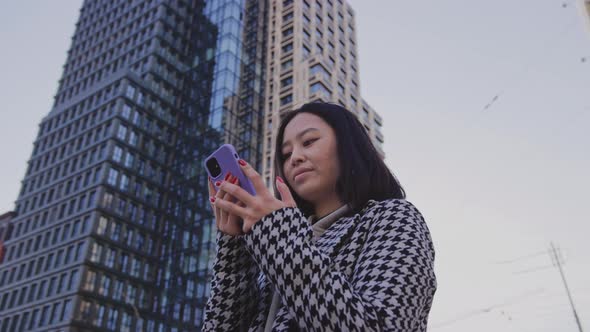 Young Adult Woman Browsing on Her Smart Phone in City Downtown Beneath a Skyscraper