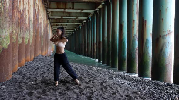 a Barefoot Woman Dances Under the Pillars of the Bridge Against the Background of the Incoming Waves