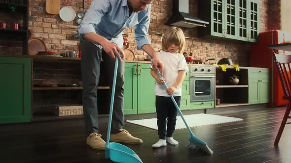 Young Dad and His Son in Casual Clothes are Sweeping the Floor and Brushing Litter Into the Scoop at