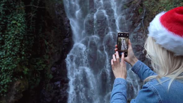 blonde in Santa Claus hat stands waterfall takes pictures photos on smartphone