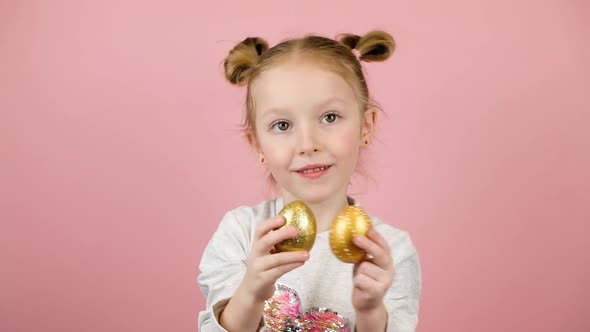 Funny Little Blonde Girl Smiling and Playing with Golden Easter Eggs on Pink Background