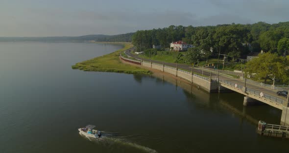 Backwards Aerial Pan of Speedboat Passing on Bay Near a Small Bridge and Forest