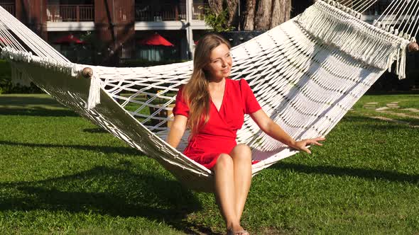Young Attractive Smiling Woman Sitting and Relaxing in Hammock in Modern Resort