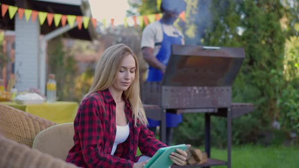 Young Slim Blond Caucasian Woman Surfing Internet on Tablet As Blurred African American Man Cooking