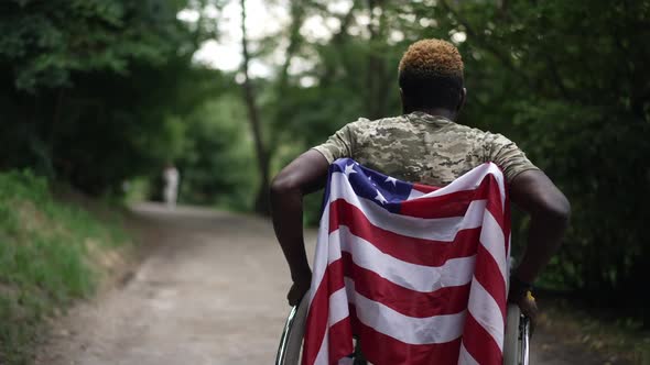 Tracking Shot African American Young Man in Wheelchair with US Flag Riding on Alley in Summer Park