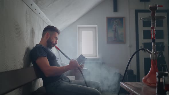 The Guy Smokes a Hookah Indoors and Exhales Smoke