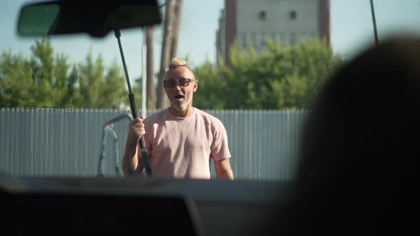 Shooting From Inside Automobile of Satisfied Caucasian Tattooed Man with High Pressure Washer