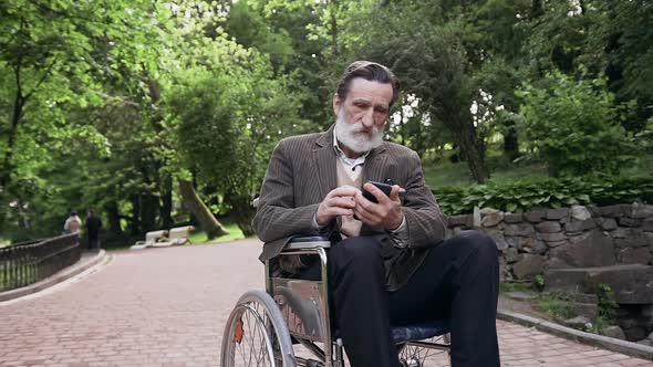 Bearded Senior Man Sitting in Wheelchair in the Middle of Urban Park and Using Phone