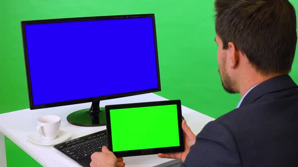 A Businessman Sits at a Desk with a Blue Computer Screen and Looks at a Tablet - Green Screen