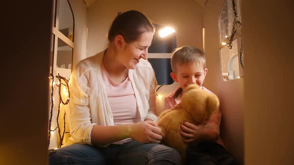 Happy Smiling Little Boy with Young Mother Playing with Toy Teddy Bear in Tent or Toy House at Night