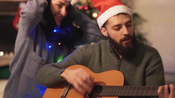 Man Playing Guitar and Singing Sitting on the Floor Near Christmas Tree in Modern Room. Happy Couple