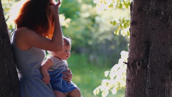 baby and mother are playing standing in the forest by a tree trunk in backlight