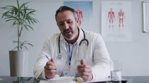 Caucasian male doctor at desk talking and gesturing during video call consultation