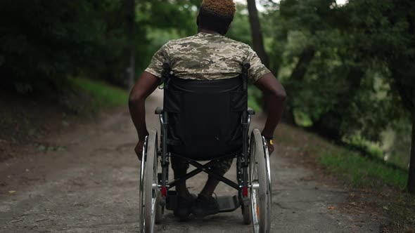 Back View African American Ill Soldier in Wheelchair Rolling Wheels with Hands Riding in Summer Park