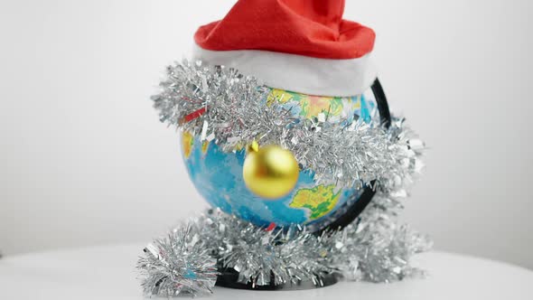 Closeup Earth Globe in Christmas Hat and Decorative Garland with New Year Balls Falling on Table in