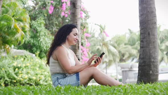 Cute Woman Use of Digital Tablet or Phone in the Street in the Park Sitting on Grass or Lawn.