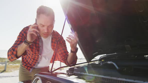 Stressed caucasian man talking on smartphone by broken down car with open bonnet on sunny day