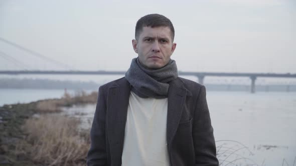 Portrait of Unhappy Caucasian Man Posing at the Background of River and City Bridge. Upset Young Guy