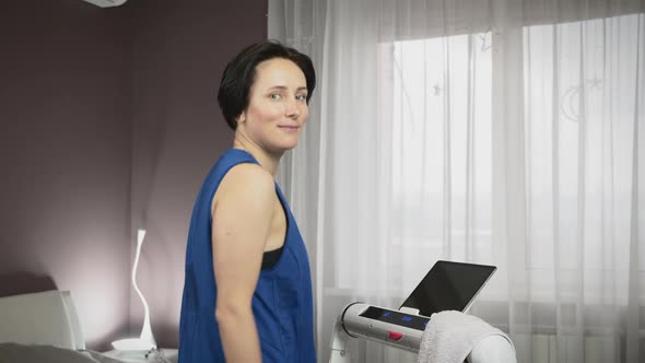 Young female is walking on treadmill, looking at camera and smiling