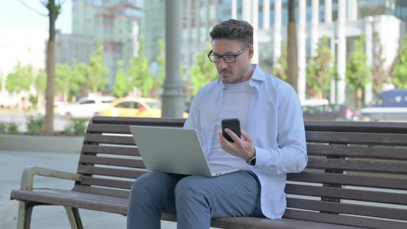 Man Using Smartphone and Laptop While Sitting Outdoor on Bench