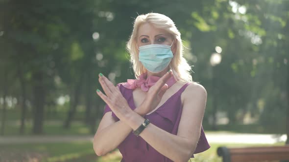 Serious Woman in Covid Face Mask Crossing Hands Looking at Camera Standing in Sunbeam Outdoors