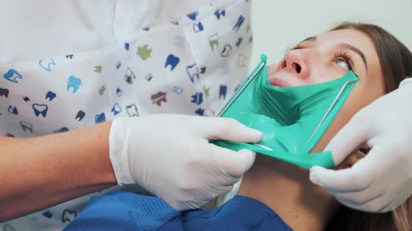 A dentist in white latex gloves makes a green rubber dam with a metal frame.