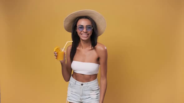 African Woman on Vacation Wearing Bikini and Hat Drinking Cocktail Over Isolated Yellow Background