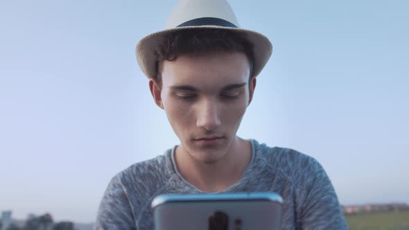 Boy in a Hat Reading Messages on His Cell Phone Blue Sky Background Camera Tracking