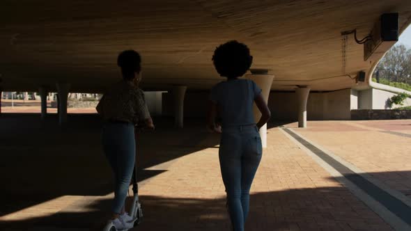 Two mixed race women riding electric scooter under bridge