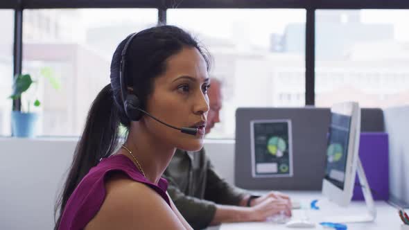 Mixed race businesswoman sitting using computer talking on phone headset in office