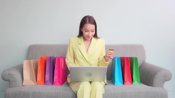 Young asian woman sitting on sofa using laptop computer shopping online with credit card buying to i