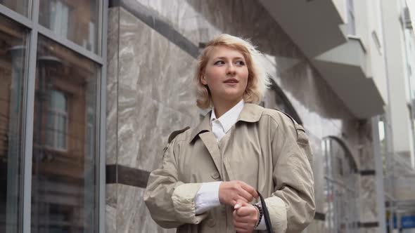 Young Blonde Woman in a Beige Trench Coat and White Blouse Stands on the Background of a Modern