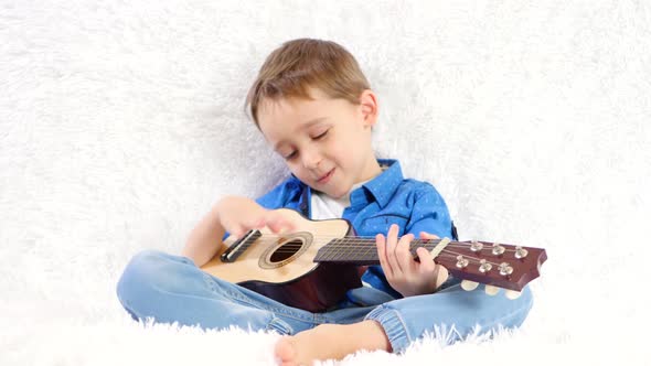 A Child with Pleasure Plays an Acoustic Guitar Sings a Song Sitting on a White Sofa