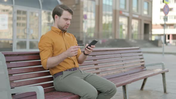 Excited Young Man Shopping Online via Smartphone, Sitting Outdoor
