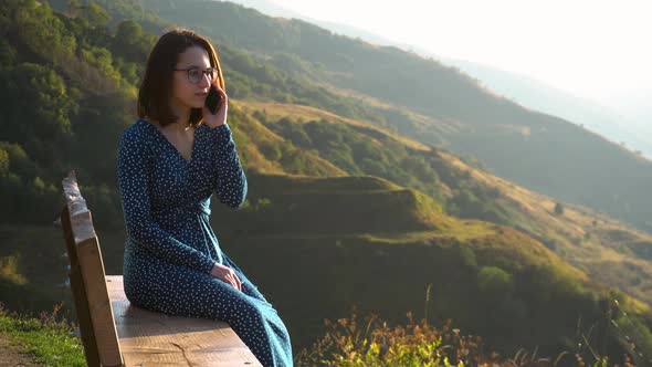 A Young Woman in a Dress Sits on a Bench and Speaks on the Phone Against a Background of Mountains