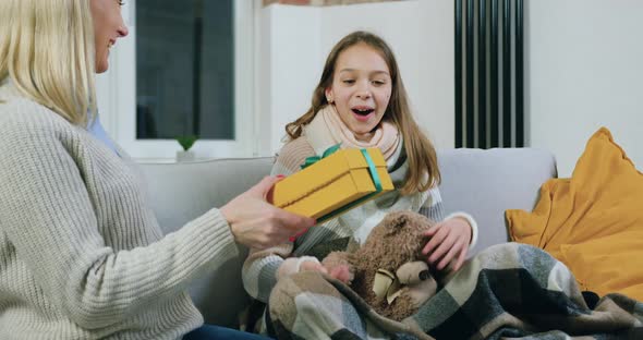 Mother Giving Gift Box to Her Surprised Cheerful Teen Daughter which Sitting on the Comfortable Sofa