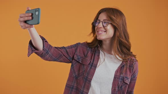 Ginger Girl in Glasses Enjoying Video Call on Smartphone Smiling Showing Victory Sign Sending Air