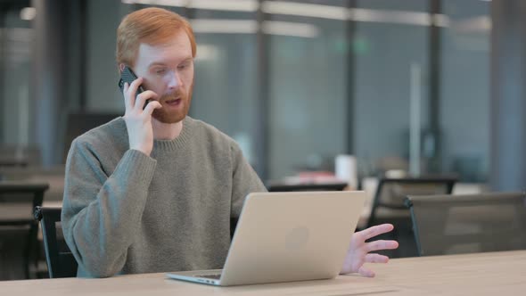 Young Man Talking on Smartphone While Using Laptop in Office
