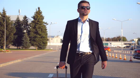 Young Successful Businessman in Sunglasses Goes with Luggage To Airport Terminal at Sunset